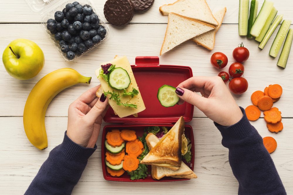 Preparing healthy vegetarian snacks on white rustic wood. Female hands making sandwiches and putting into take away plastic lunch box, top view. 