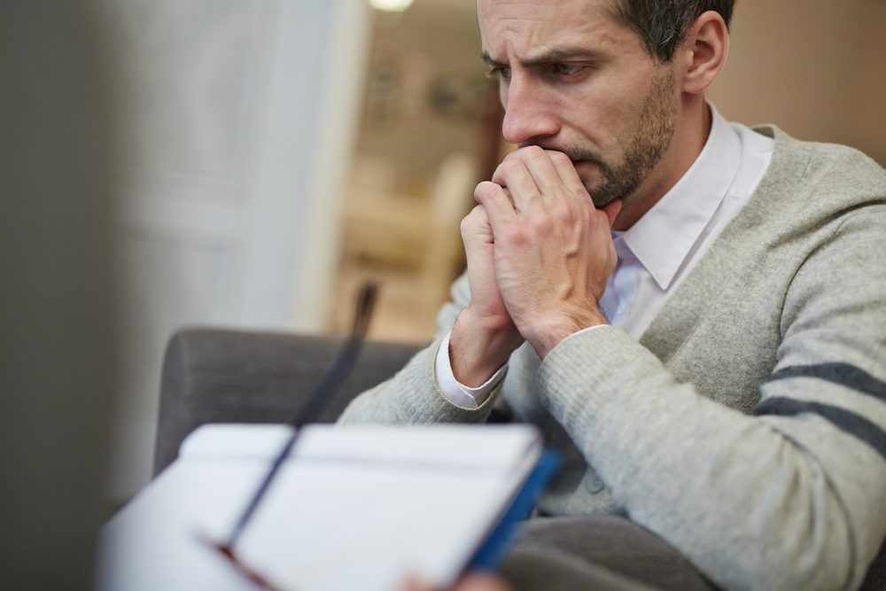 Waist-up portrait of disappointed bearded patient absorbed in thoughts while his psychiatrist filling in medical card