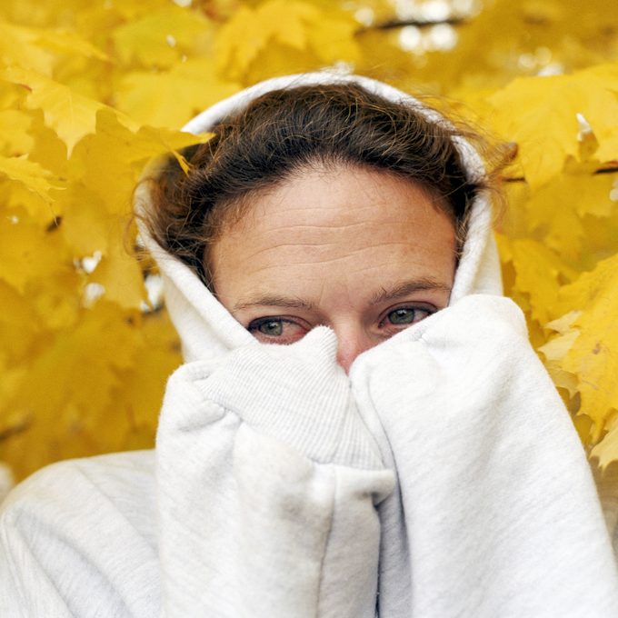 Woman wearing hooded sweatshirt pulled over her face with yellow autumn leaves behind her