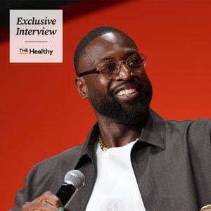A smiling man with a beard and glasses holds a microphone, wearing a gray jacket and white shirt, in front of a red background. Text reads: "Exclusive Interview, The Healthy.