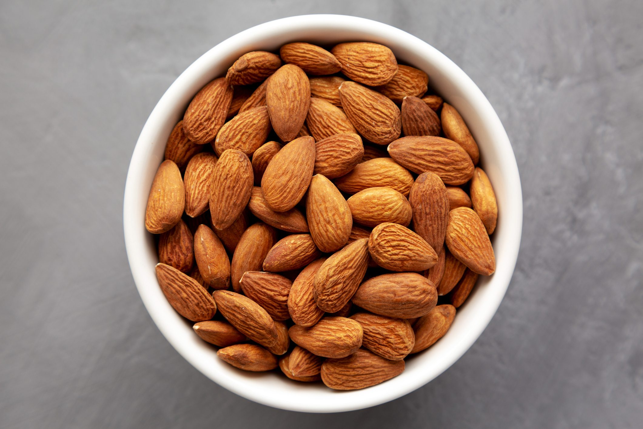 Raw brown almonds in a bowl on a gray surface, top view. Flat lay, overhead, from above.