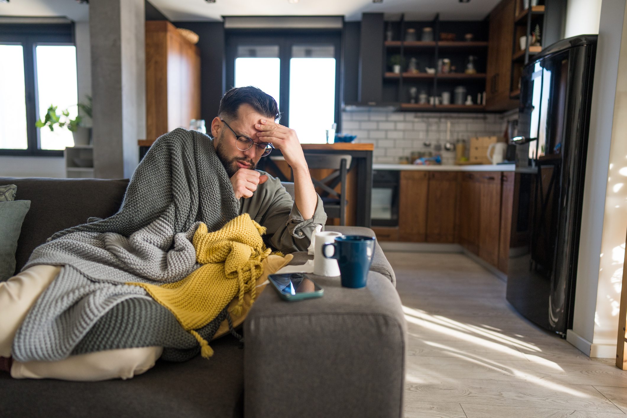 Sick man coughing at home wrapped in blankets on sofa